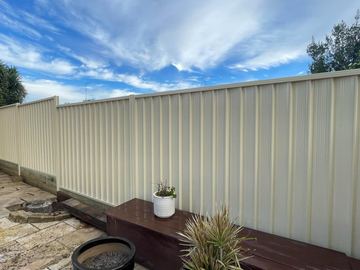 Cream Colorbond privacy fence along a paved courtyard with a low outdoor bench and potted plants