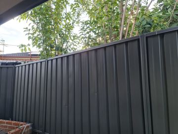 Monument corrugated panels stand before slender green trees with a red brick planter edge in the foreground.