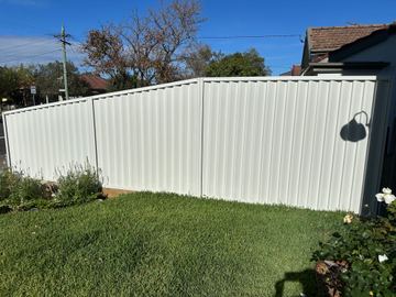 The scene shows a light grey corrugated steel fence where garden beds sit at the base.