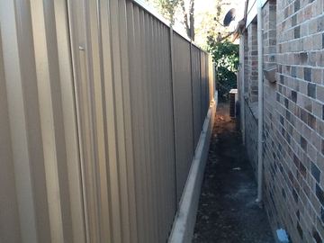 Cream Colorbond privacy fence tight against a brick side wall with a narrow dirt gap