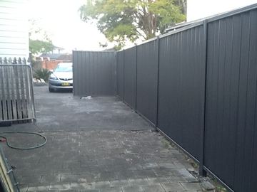 Charcoal Colorbond privacy fence enclosing an empty paved courtyard with a garden hose on the ground