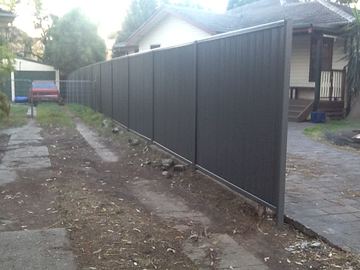 Long charcoal Colorbond privacy fence stretching across a dirt yard toward a weatherboard house