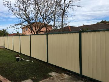 Cream Colorbond privacy fence with dark posts across a lawn, with a bucket on the grass