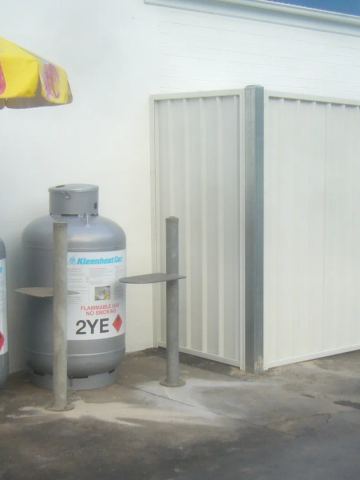 A cream corrugated steel enclosure with a wide gate shields equipment next to Kleenheat gas bottles on an asphalt forecourt.