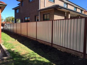 Cream Colorbond privacy fence with brown posts on a timber plinth beside a two-storey brick house