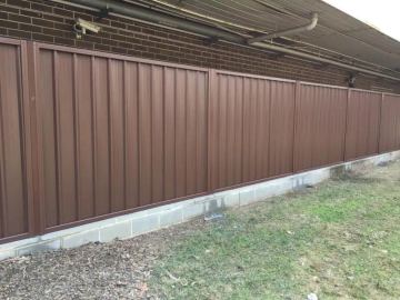 Warm jasper-brown panels mount on a row of grey concrete sleepers along a brick wall with patchy grass at the base.