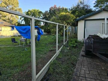 Cream skeleton fencing with sleepers runs beside a cream shed, trailer and tarp-covered chairs on grass.