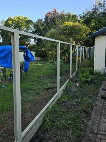 Cream skeleton fencing with sleepers runs beside a cream shed, trailer and tarp-covered chairs on grass.