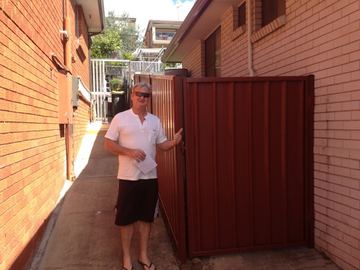 Manor-red corrugated gate and panel close a narrow concrete alley between orange and buff brick walls.