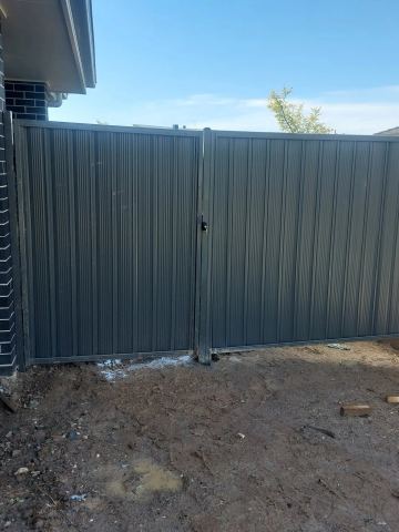 Charcoal double swing gates latch on muddy ground beside a dark brick wall with a shovel and packaging nearby.