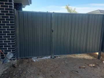 Charcoal double swing gates latch on muddy ground beside a dark brick wall with a shovel and packaging nearby.