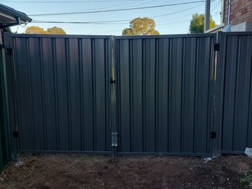 Charcoal double gates with drop bolts stand on fresh soil beside a red brick wall and overhead wires.