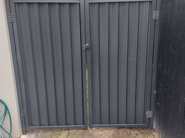 Charcoal double gates hinge between a rendered wall and a black timber paling fence on old brick pavers.