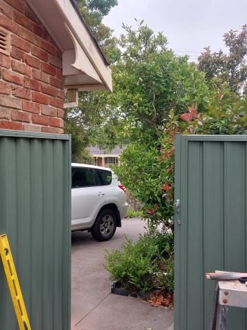 Pale-green corrugated panels with posts flank a brick house driveway where a white SUV is parked and tools sit on a stool.