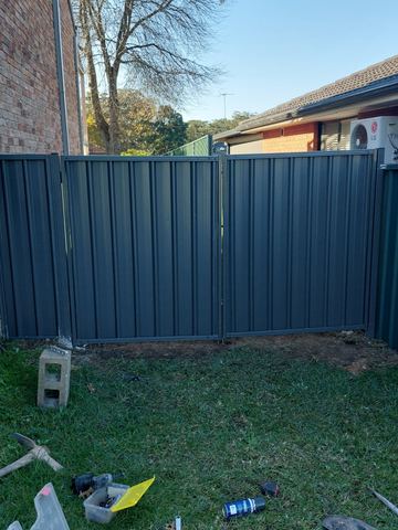 Wide charcoal double gates span between brick walls with solar gear on one side and an air conditioner on the other.