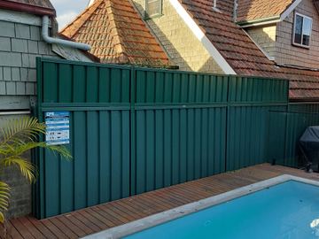 Deep green tall panels with a resuscitation sign shield a timber pool deck beside glass pool fencing.