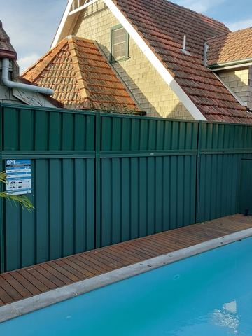 Deep green tall panels with a resuscitation sign shield a timber pool deck beside glass pool fencing.