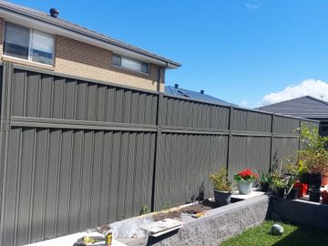 Charcoal panels mount on split-face grey blocks above lawn with potted geraniums and tools on a board.