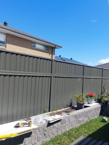 Charcoal panels mount on split-face grey blocks above lawn with potted geraniums and tools on a board.