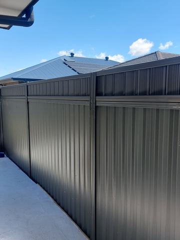 Charcoal panels with a small upper inset line a light concrete path beside dark brick and a neighbour's solar roof.