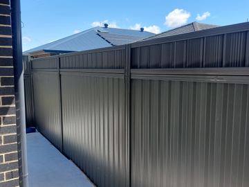 Charcoal panels with a small upper inset line a light concrete path beside dark brick and a neighbour's solar roof.