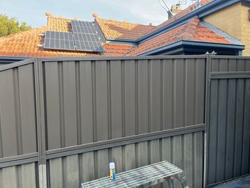 Charcoal lower panels with basalt upper sheets include a narrow ribbed gate beside a terracotta roof with solar panels.
