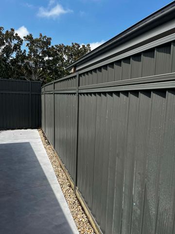 Charcoal panels on sleepers meet a concrete corner with tan pebbles and magnolia trees above the fence line.