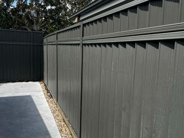 Charcoal panels on sleepers meet a concrete corner with tan pebbles and magnolia trees above the fence line.