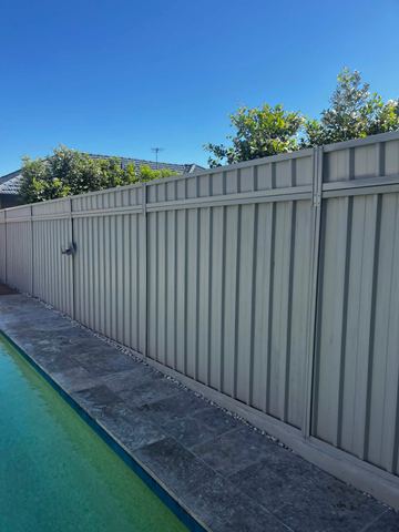 Cream panels and a latched gate run beside travertine pool coping and white pebbles with trees overhead.