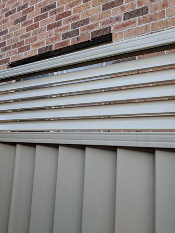 Cream Colorbond fence with a white slat topper bolted against a brick house wall