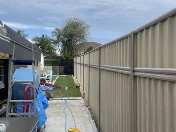 Cream Colorbond privacy fence along a concrete backyard path with an installer at work