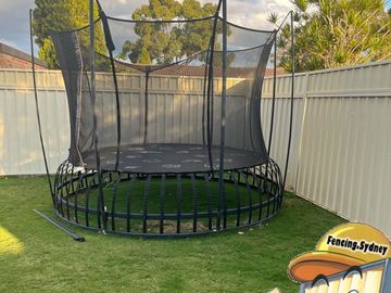 Cream panels wrap a lawn corner behind a large trampoline with suburban trees and sky above.