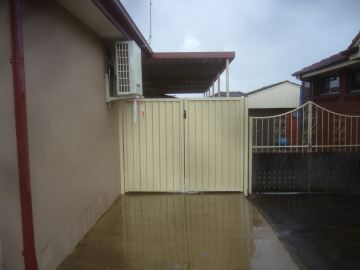 Cream Colorbond double gate under a carport on a wet concrete driveway