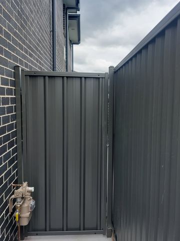 Charcoal gate and tall panel attach to dark brick beside a gas meter on a paved side access.