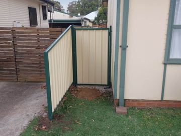 Cream gate and panel in green frames create a small enclosure between a cream house wall and a timber slat fence.