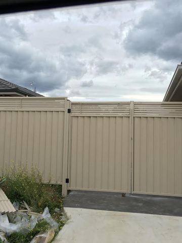 Cream panels and louvre-topped double gates span a driveway beside a red brick garage under grey clouds.