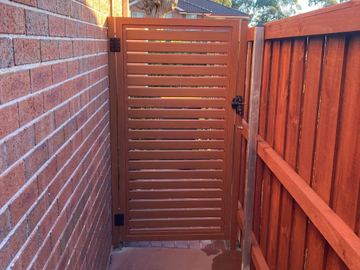 Red-brown horizontal slat gate opens on a tinted concrete path between brick and matching timber-look fencing.