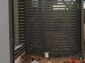 Charcoal slat gate and fixed panel bolt to dark brick on rocky soil with an electrical box behind.
