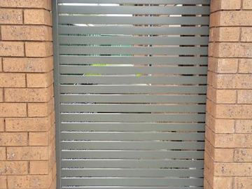 Taupe horizontal slats fill a custom curved gate within a brick arch over concrete and a drainage grate.