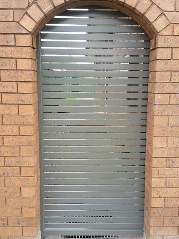 Taupe horizontal slats fill a custom curved gate within a brick arch over concrete and a drainage grate.