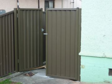 The scene shows a mid grey corrugated steel fence where a gate sits in the run, concrete paving runs alongside.