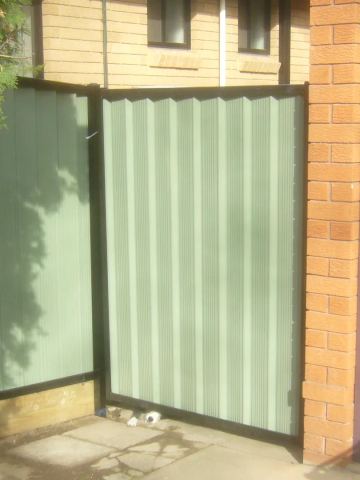 The scene shows a green corrugated steel fence where a gate sits in the run, sleepers form the base.