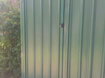 The scene shows a mid grey corrugated steel fence where a gate sits in the run, concrete paving runs alongside.