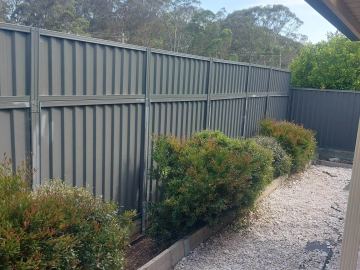 Olive panels on timber-edged beds with red-tipped shrubs sit above white pebbles where a spirit level leans against the fence.
