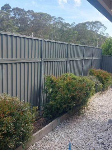 Olive panels on timber-edged beds with red-tipped shrubs sit above white pebbles where a spirit level leans against the fence.