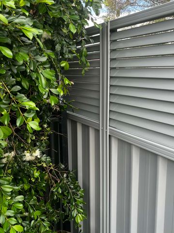 Shale-grey corrugated sheets with a louvre topper meet a leafy hedge beside a black tubular fence corner.