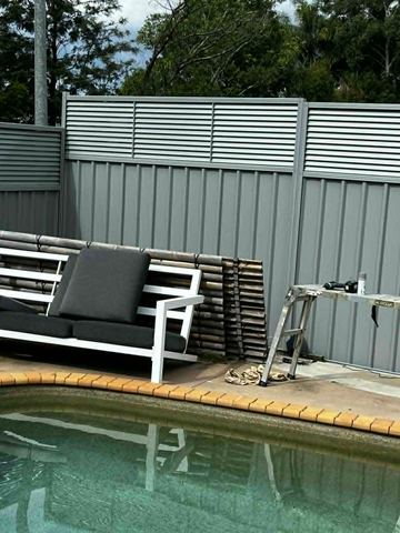 Basalt-grey panels with a louvre top border a pool deck where outdoor seating and a ladder sit on terracotta coping.