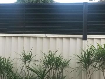 The scene shows a mid grey corrugated steel fence where louvre sections add ventilation, garden beds sit at the base.