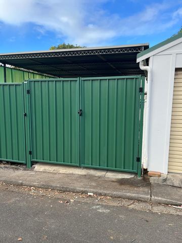 Green corrugated double gates hang beside a white garage wall opening onto asphalt with a metal carport roof behind.