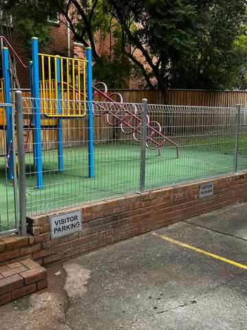 Silver weldmesh with rolled edges caps a brick retaining wall around synthetic turf, a yellow slide and visitor parking signs.